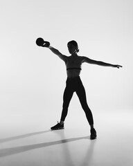 Black and white photography. Full-length image of young woman with slim body, training, doing exercises with kettlebell. Concept of sport, active and healthy lifestyle, workout, fitness