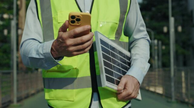 Man construction worker holds smartphone and solar panel walking along bridge in evening. Male professional in safety vest examines project details on phone before night shift