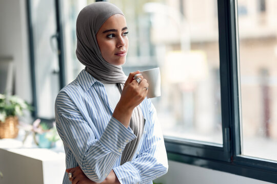 Young woman holding coffee cup and looking through window at home