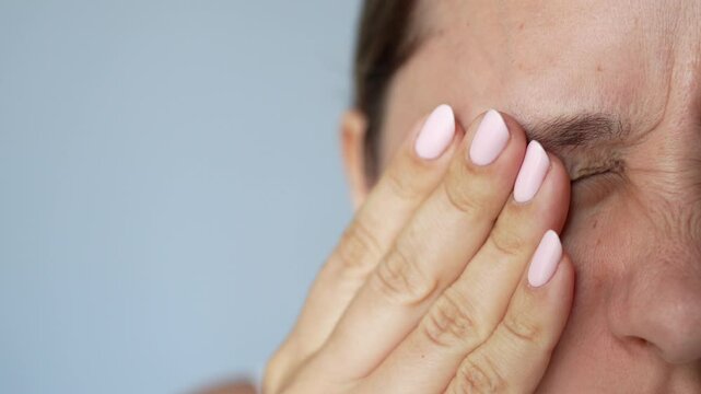 Young caucasian blonde woman covering her eyes pressing on them with hand suffering from eye pain on gray background. Photophobia, painful sensitivity to bright light. Ophthalmology concept