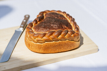 Ein herzförmiges Brot mit geflochtenem Rand liegt auf einem Holzbrett, daneben ein Messer. Die Szene ist schlicht und zeigt das Brot im Sonnenlicht.