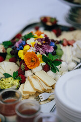 Eine stilvolle Käseplatte, dekoriert mit essbaren Blüten, frischem Obst wie Trauben, Erdbeeren und Himbeeren sowie Kräutern. Neben der Platte befinden sich Schalen mit Saucen und Dips. 