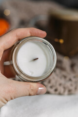 A woman's hand holds a candle with a wooden wick against a background of lights and a knitted plaid. Vertical photo