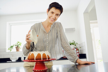 Smiling woman pouring cream on cake in kitchen at home