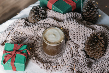 Winter still life. A glass cup of coffee with milk, big cones, lights, a green gift box with a red bow. Modern christmas rustic eco friendly decor, scandinavian winter hygge. Atmospheric christmas