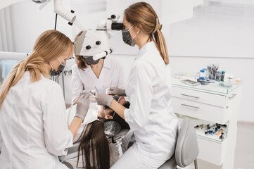 Obraz premium Female doctor dentist and two assistants treating teenage girl's teeth under microscope. team of dentists, teamwork in the clinic. stomatologist examining the mouth of a patient lying on the chair