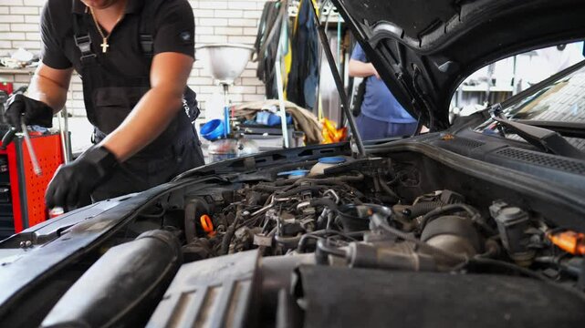Young repairman working under hood of vehicle at service. Professional auto mechanic fixes engine of car using screwdriver tool at workshop. Male repairer checking automobile motor at garage. Slow mo