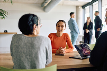 A diverse team of business professionals engaged in a discussion around a conference table in an office, while their colleagues collaborate in the background