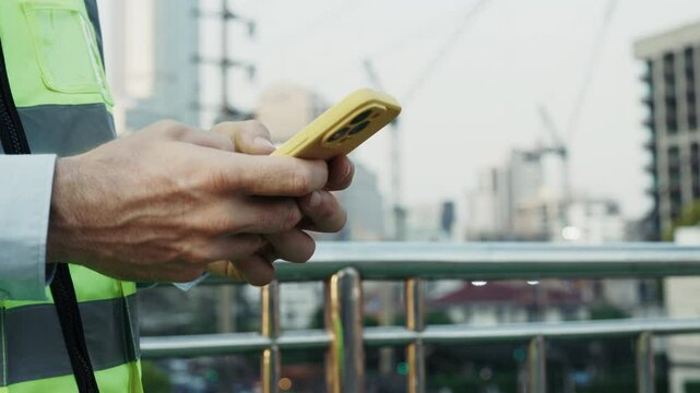 Man engineer in safety vest types message on smartphone. Male contractor texts team members coordinating tasks and providing instructions for next steps on construction site. Close up