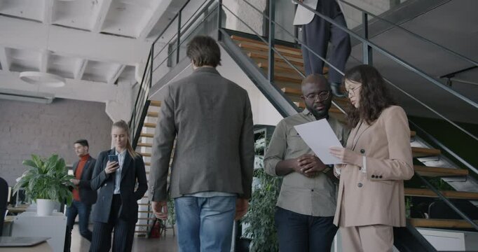 Two businesspeople discussing document standing in office while colleagues walking around. Professional collaboration and workplace concept.