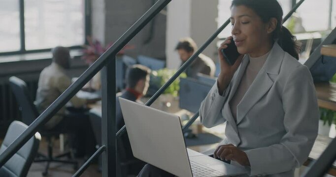 Middle Eastern business lady speaking on mobile phone and using laptop working on stairs in open space office. Businessperson and communication concept.