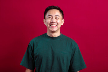 A cheerful young Asian man in a green T-shirt smiles warmly, standing confidently against a bold red background, exuding positivity and approachability in a relaxed and vibrant setting