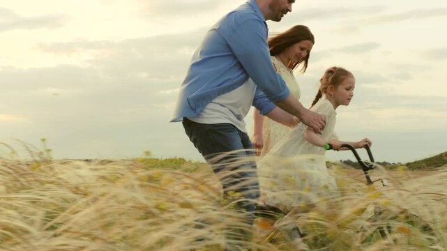 happy parents teach smiling child daughter ride bike. happy family concept. childhood dream ride bike. kid turns handlebars bike. children fast cycling. caring father mother kid daughter park meadow