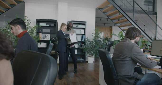 Zoom-in of man and woman colleagues standing in office talking discussing documents while people working at desks using computers. Job and coworking concept.