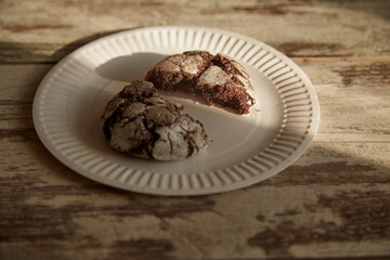 A plate with a broken half of a chocolate cookie with cracks on an old rustic wooden table in a beam of sunlight, close-up, homemade baking