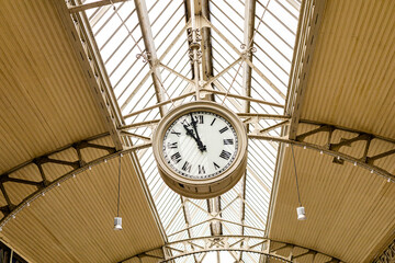 a large clock suspended from the ceiling of a majestic building with a glass roof. The clock...