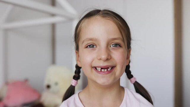 Close-up of smiling Caucasian girl with brown hair and braided pigtails, showing gaps from missing baby teeth, in a bright home setting. - Powered by Adobe