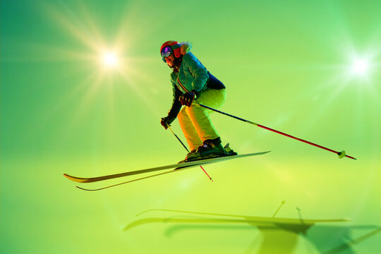 Action shot of skier leaping forward, as she jump over obstacles vibrant motion against green studio background with sunbeams. Concept of extreme sport, Christmas holidays. Ad