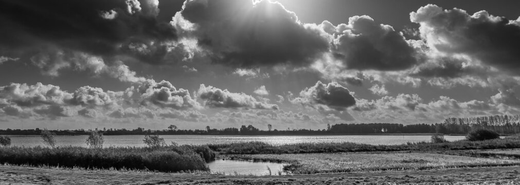 Dark clouds gather dramatically over a serene Dutch landscape, casting shadows across a large body of water and distant trees. Sunlight breaks through in places, creating a striking contrast.