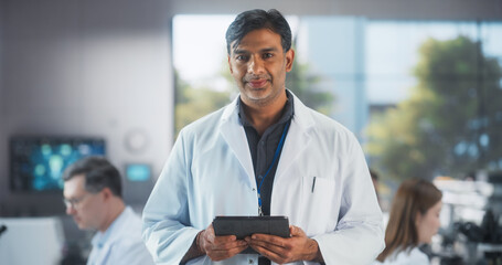 Portrait of an Indian Adult Male Scientist Using Tablet Computer, Looking at Camera and Smiling in...