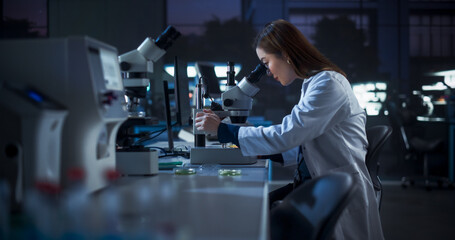 Obraz premium Medical Research and Development Laboratory: Hard-Working Asian Female Scientist Using Computer and Looking at a Sample in a Petri Dish Under a Microscope. Advanced Genetic Engineering Lab at Night