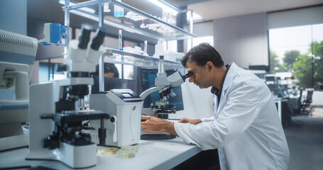 Genetic Research and Development Laboratory: Adult Scientist Working on a Computer and Looking at a Sample in a Petri Dish Under a Microscope. Advanced Biotechnology Lab with Modern Equipment