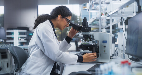 Close Up Portrait of a Latin Female Microbiologist Wearing Safety Glasses and Looking Through the Microscope, Analyzing Sample Cells. Skillful Scientist Working in a Medical Science Laboratory