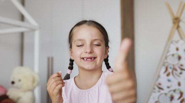 Happy Caucasian girl with braided hair showing fallen baby tooth in cozy home setting, expressing excitement.
