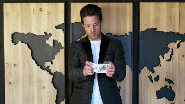 Stylish magician wears suit, is performing a card trick in front of a world map indoors at studio.