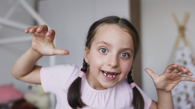 Portrait of playful little Caucasian girl with braided hair and gaps from missing milk teeth posing at home with frightening expression.