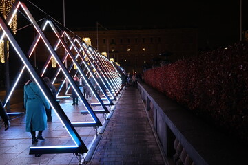 Futuristic light installation with illuminated triangular frames and people walking through a vibrant pathway at night.