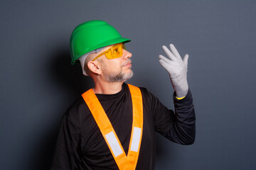 Photo of a civil worker wearing a green helmet, reflective vest and protective glasses, showing his fingers. Isolated on gray background.