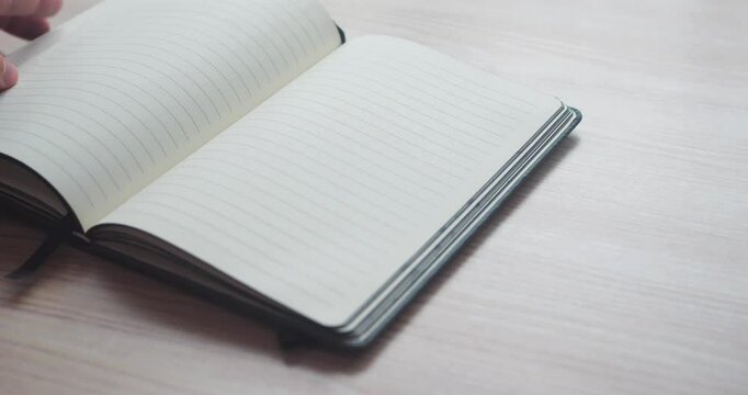 Rack focus close-up of a man opening a minimalist notebook on a wooden table and writing down ideas or notes. Man writing an inscription in a notebook. 4K