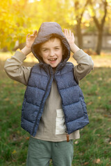 Young child in a gray puffer vest and beige long-sleeve shirt in a park with autumn foliage. The...