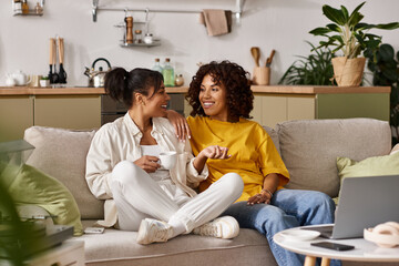 Two young women share laughter and coffee while chatting in their stylish apartment.
