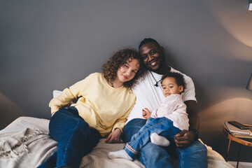 Portrait of biracial family enjoying together time spending in home interior, Caucasian woman and African American smiling at camera during positive resting leisure with multicultural child
