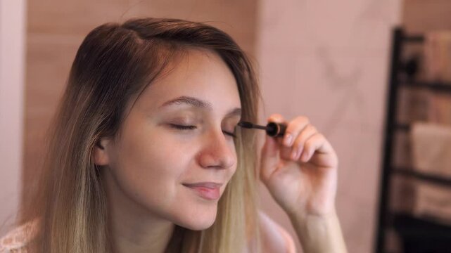 blonde girl paints her eyelashes with mascara at home in front of the mirror. close up. Selective focus