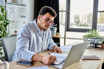 A focused man writes notes while using a laptop in a well lit office space.