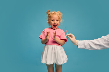 Happy little blonde girl, child smiling, clapping hands while doctor making vaccine injection against blue studio background. Concept of healthcare, medical treatment, vaccination, childhood