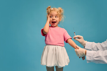 Emotive little girl, child in pink t-shirt holding hand on head, with wide open eyes and mouth, while doctor making vaccination against blue studio background. Concept of healthcare, medical treatment