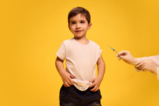 Little boy, child in black and white clothes calmly standing while doctor making vaccine injection with syringe against yellow studio background. Concept of healthcare, medical treatment, vaccination
