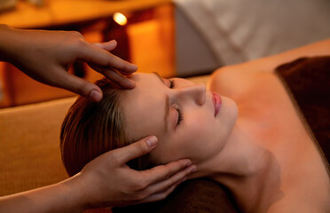 Caucasian woman enjoying relaxing anti-stress head massage and pampering facial beauty skin recreation leisure in warm candle lighting ambient salon spa in luxury resort or hotel. Quiescent