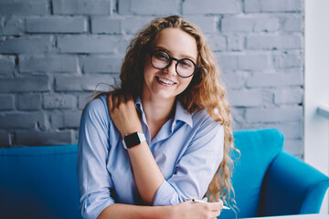 Portrait of happy hipster girl in trendy spectacles for provide eyes protection sitting in cafeteria and smiling at camera during free time on leisure, best seller or textbook staying on table