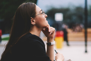 Positive female writer with textbook for education thinking about romantic article for content blog, successful smiling hipster girl enjoying free time after preparing for university test outdoors