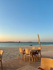 Serene beach at sunset. Wicker chairs and tables on tiled patio. Sandy shore leading to calm water. Red and yellow flags. Clear sky with gradient from light blue to warm hues. Peaceful atmosphere. 