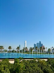 Paved park path with green hedges and palm trees. Curved metal railing along path. Clear blue sky. Urban skyline with tall buildings in the background. Person walking on the path.