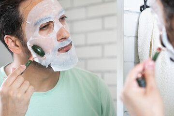 Handsome man affixes collagen anti-aging face sheet mask in bathroom. Skin care and Men's Grooming concept