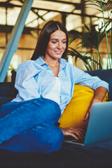 Positive smiling hipster girl searching comedy show for watching using modern laptop device indoors, happy woman resting on comfortable sofa while messaging with friends via application on netbook