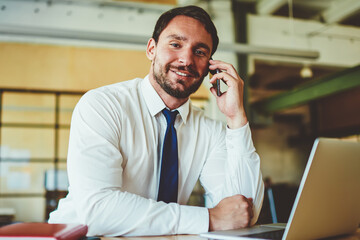 Portrait of successful businessman looking at camera and enjoying friendly telephone conversation during work process with computer in modern coworking space, positive man calling via smartphone