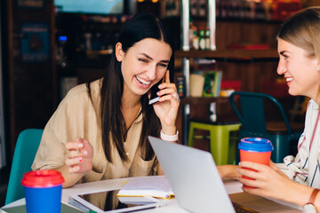Cheerful woman using mobile application for making business conversation during cooperation remote meeting with smiling colleague, emotional colleagues laughing during brainstorming in coffee shop
